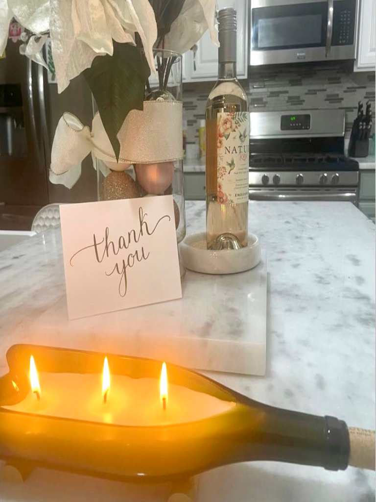Green glass bottle repurposed as a candle holder with three lit candles inside with a thank you card, a jar of flowers, and bottle of white wine. 