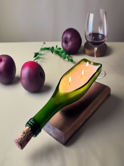 A upcycled green wine bottle candle with three wicks burning, placed on a wooden surface, with plums and a glass of red wine in the background.