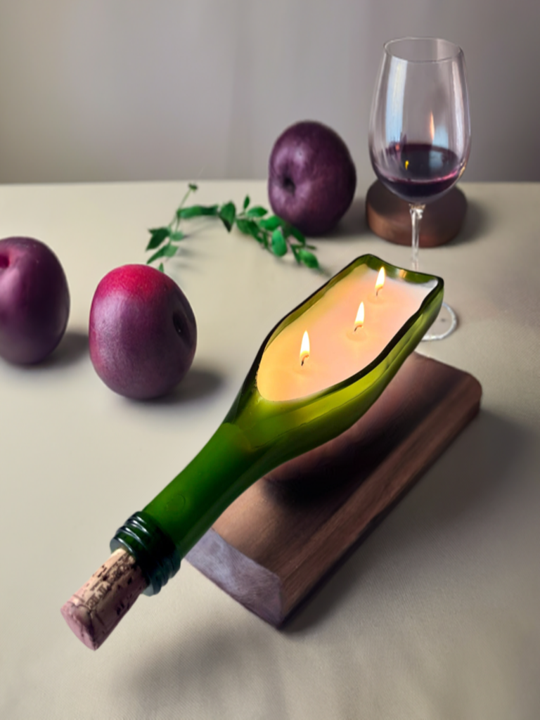 A upcycled green wine bottle candle with three wicks burning, placed on a wooden surface, with plums and a glass of red wine in the background.